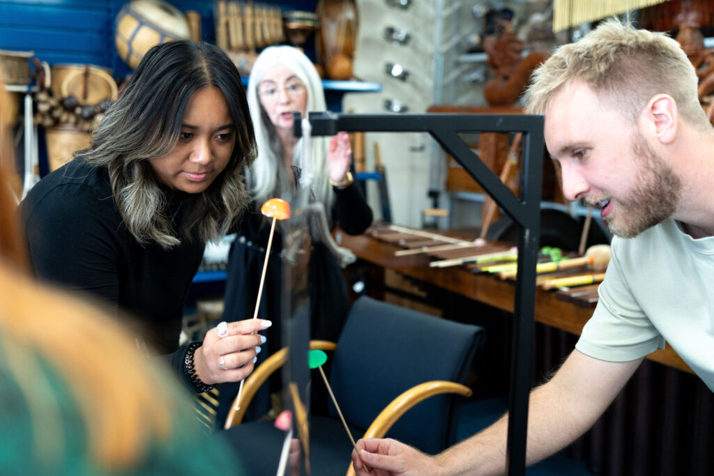 A young woman and a young man, who are participating in a 'Who's Listening' workshop. They are attempting to play a Thundersheet, as Evelyn looks on (slightly blurred in the background).