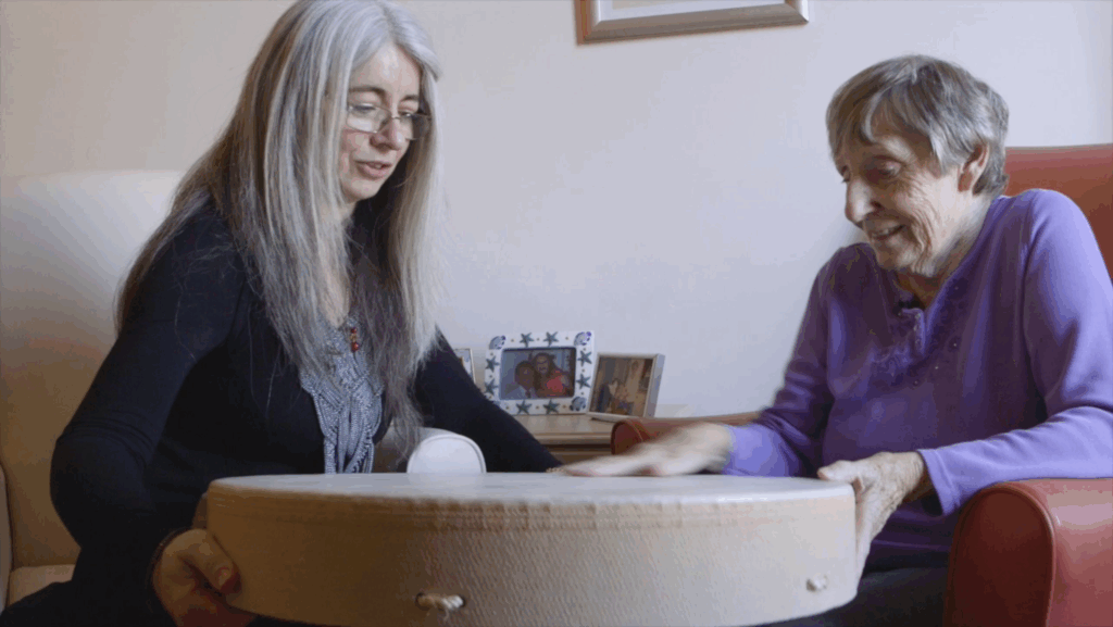 Dame Evelyn and Babs, an elderly white woman with short grey hair, wearing a purple sweater. They are seated facing each other in room at the care home. Between them sits a large frame drum. Just behind are family photographs. Evelyn is encouraging Babs to interact with the Drum and listening to her thoughts. Babs is smiling and pointing at the drum as she speaks.