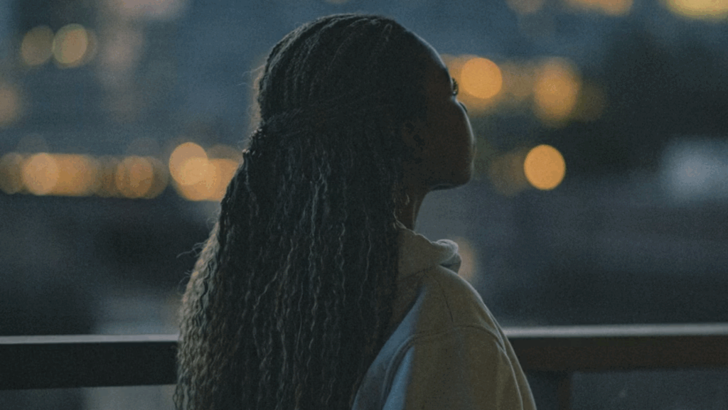 A person with long, braided hair is standing on a balcony at night, looking out over a cityscape with blurred lights in the background. The person is wearing a light-colored hoodie and is seen from behind.