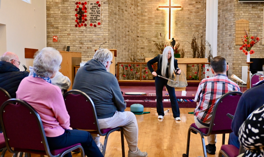 Dame Evelyn Glennie, a woman with long white hair stands at the front of a church, demonstrating a percussion instrument to a seated audience, who are listening intently. The setting includes rows of chairs, a wooden floor, and a brick wall with a large illuminated cross. Various percussion instruments are placed on the floor near the presenter, and a banner with text hangs on the wall.
