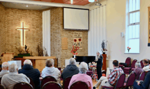 Dame Evelyn Glennie, a woman with long white hair stands at the front of a church, demonstrating a percussion instrument to a seated audience. The setting includes rows of chairs, a wooden floor, and a brick wall with a large illuminated cross.