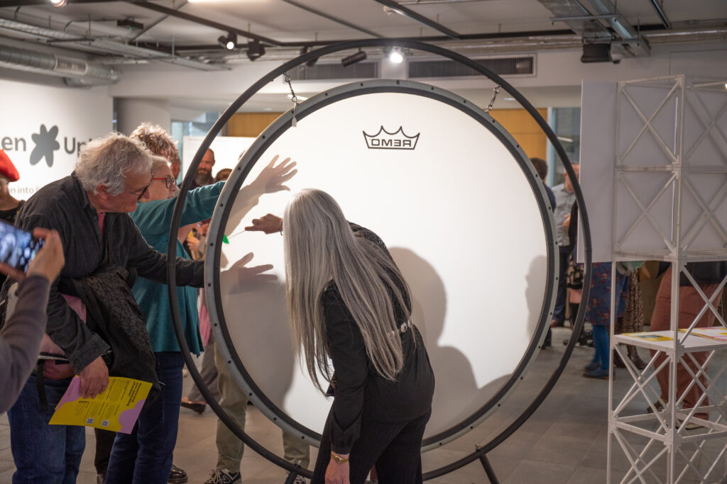 Dame Evelyn Glennie, with her back to camera, manipulpates the sound of a large gong drum with a small mallet, whilst several viewers watch on from the left of the shot, leaning in to experience the vibrations more closely.