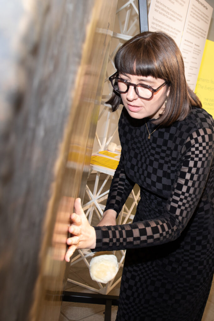 A person wearing glasses and a black checkered outfit stands beside a large hanging Tam Tam, placing one hand on its surface while holding a soft-headed mallet near the instrument. The gong fills much of the left side of the image, with exhibition panels and display materials visible in the background. The indoor setting is warmly lit, highlighting the reflective metal surface of the gong and the interaction with the instrument.