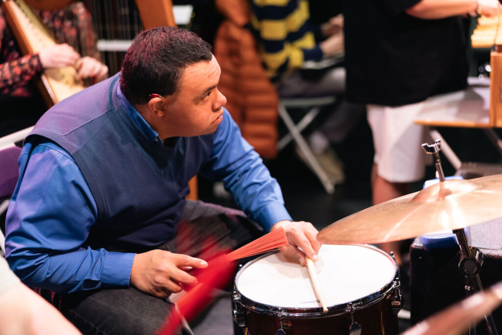 A person seated at a drum kit plays a snare drum with a drumstick in one hand and red brush sticks in the other. The person wears a blue shirt and dark blue sweater vest and has a visible hearing device. A cymbal is positioned in the foreground, while blurred musicians and instruments, including a harp, appear in the background under warm stage lighting.