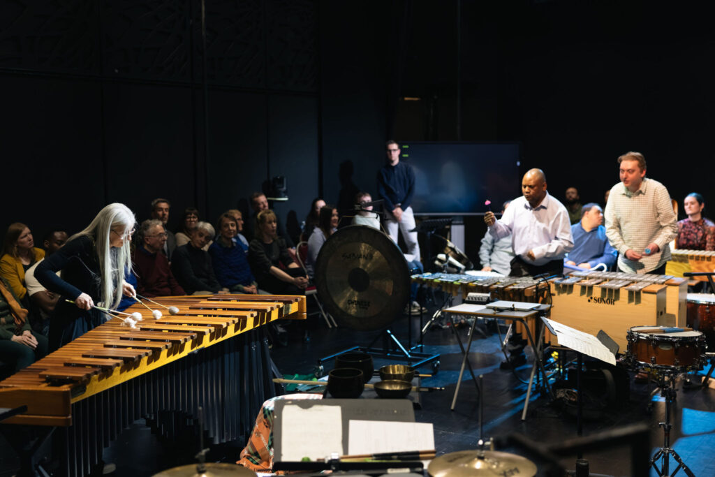 Musicians perform on a stage surrounded by an audience seated close to the performance area. In the foreground, a large marimba is being played, while other performers play percussion instruments including a vibraphone, gong, drums and singing bowls. The audience sits around the edge of the space, creating an immersive concert setting. Warm stage lighting highlights the instruments and performers against a dark background.