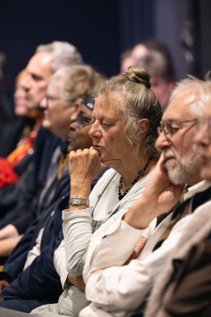 A row of audience members sits closely together during a live performance. In the centre of the image, a person with grey hair tied in a bun sits with eyes closed and a hand resting against the chin. Other attendees are seated on either side, some wearing glasses. The photograph uses a shallow depth of field, keeping the central figure in focus while the surrounding audience is softly blurred. Warm indoor lighting creates a calm and reflective atmosphere.