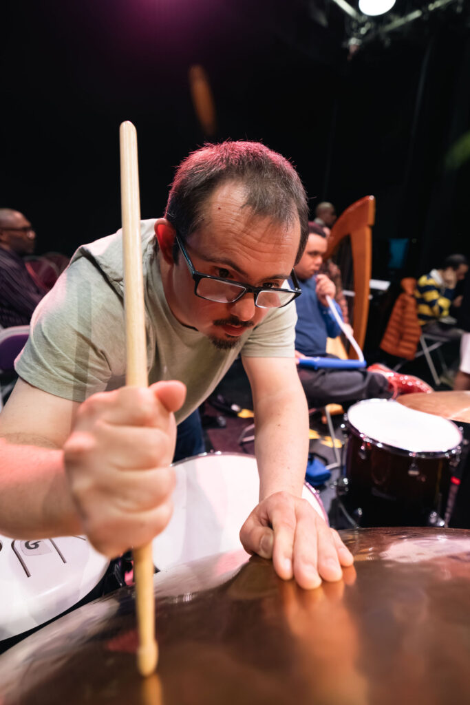 A person wearing glasses leans over a drum kit on a stage, holding a drumstick upright in one hand while resting the other hand on a cymbal. The image is taken from a low, close-up angle, with the cymbal and drumstick prominent in the foreground. Additional percussion instruments and musicians are visible but out of focus in the background, including a harp and a seated performer. Warm stage lighting highlights the scene.