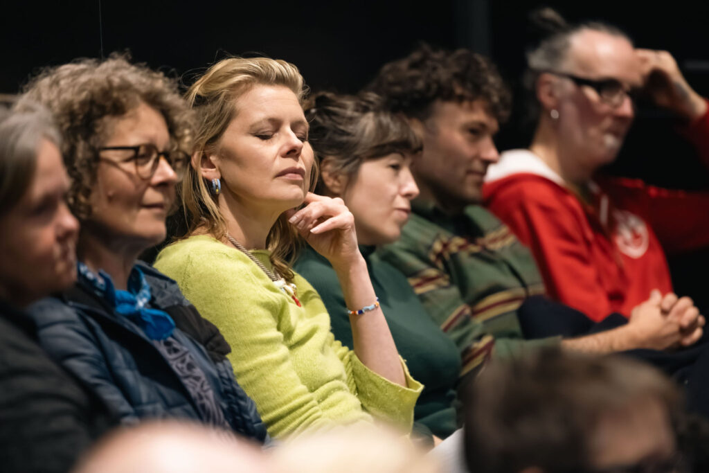 Audience members sit in a row during a live event, listening attentively. One person in a light green sweater is shown in sharp focus with eyes closed and a hand resting near the chin, while other attendees sit alongside them. The background is softly blurred, and warm indoor lighting creates an intimate atmosphere.