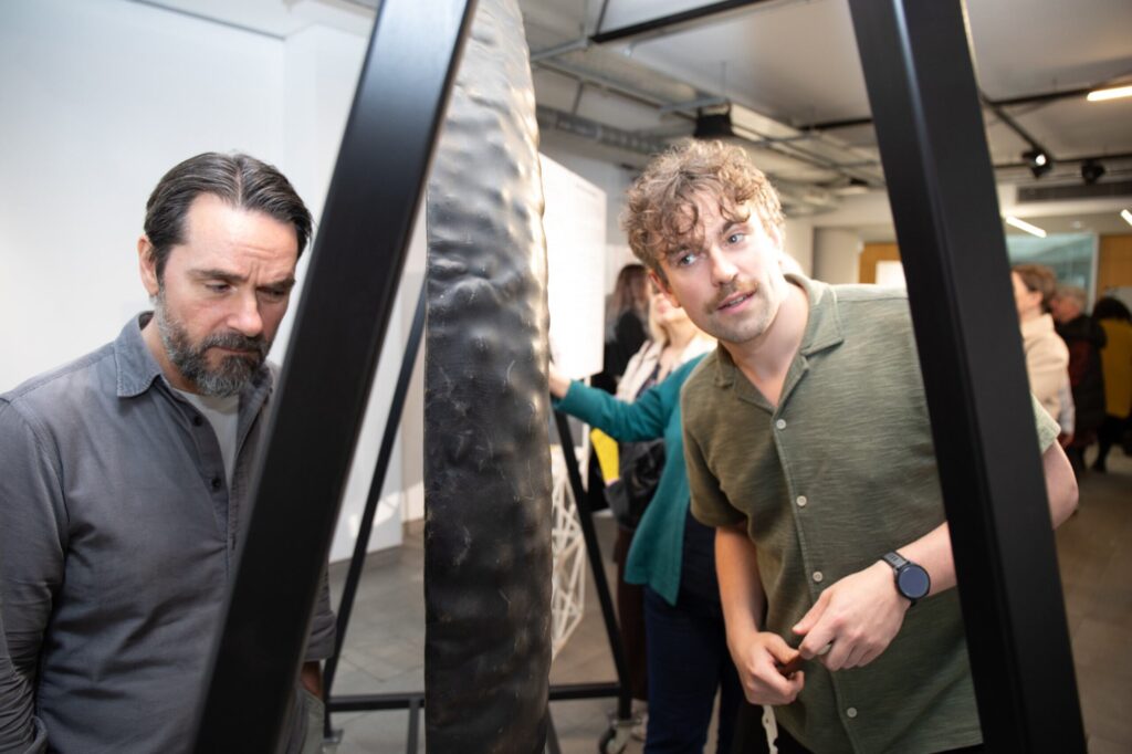 Two visitors stand beside a large suspended Tam Tam in an exhibition space, closely examining its textured metal surface. The gong is framed by black support structures in the foreground. Information panels and other visitors can be seen in the background, slightly out of focus. Bright indoor lighting illuminates the instrument and the exhibition environment, highlighting the interactive nature of the display.